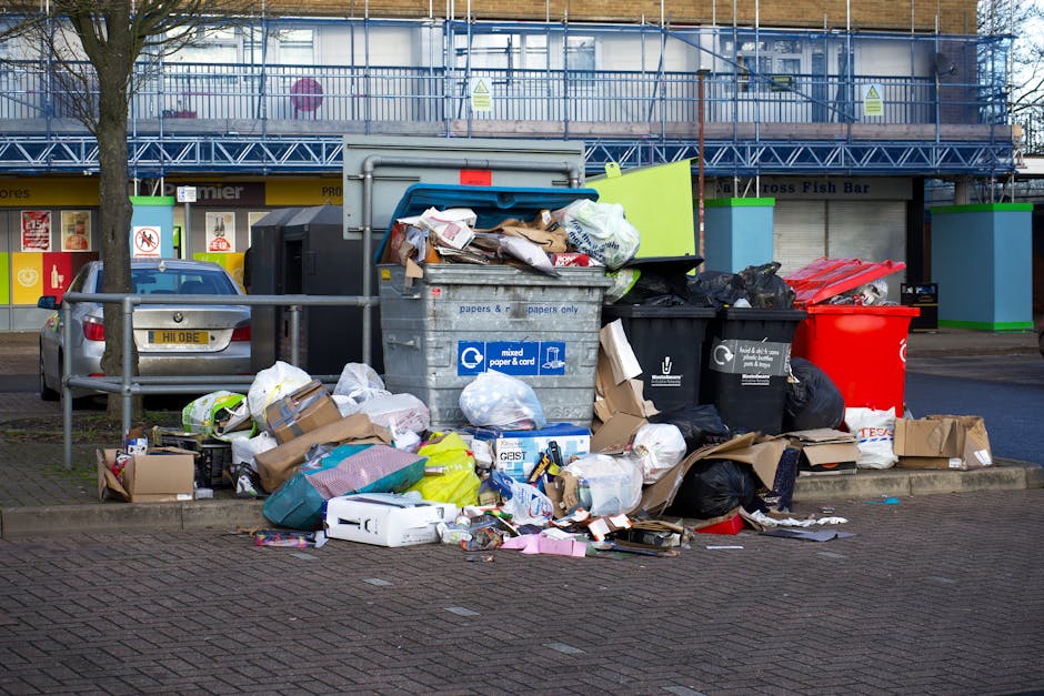 A large collection of mixed waste and rubbish, including cardboard boxes, plastic bags, and paper debris, is piled up around multiple rubbish bins in an urban parking area. The bins consist of a grey 