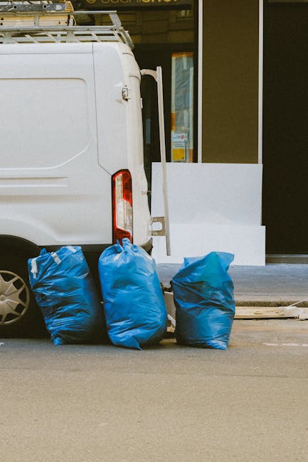 A large collection of mixed waste and rubbish, including cardboard boxes, plastic bags, and paper debris, is piled up around multiple rubbish bins in an urban parking area. The bins consist of a grey 