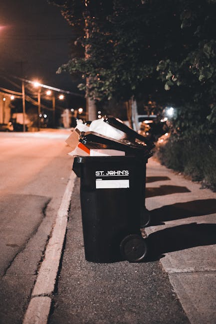 A person walking outdoors on a dirt path carrying multiple large white plastic bags filled with rubbish, with their face obscured by the bags. They are dressed in dark clothing and wearing yellow rubb
